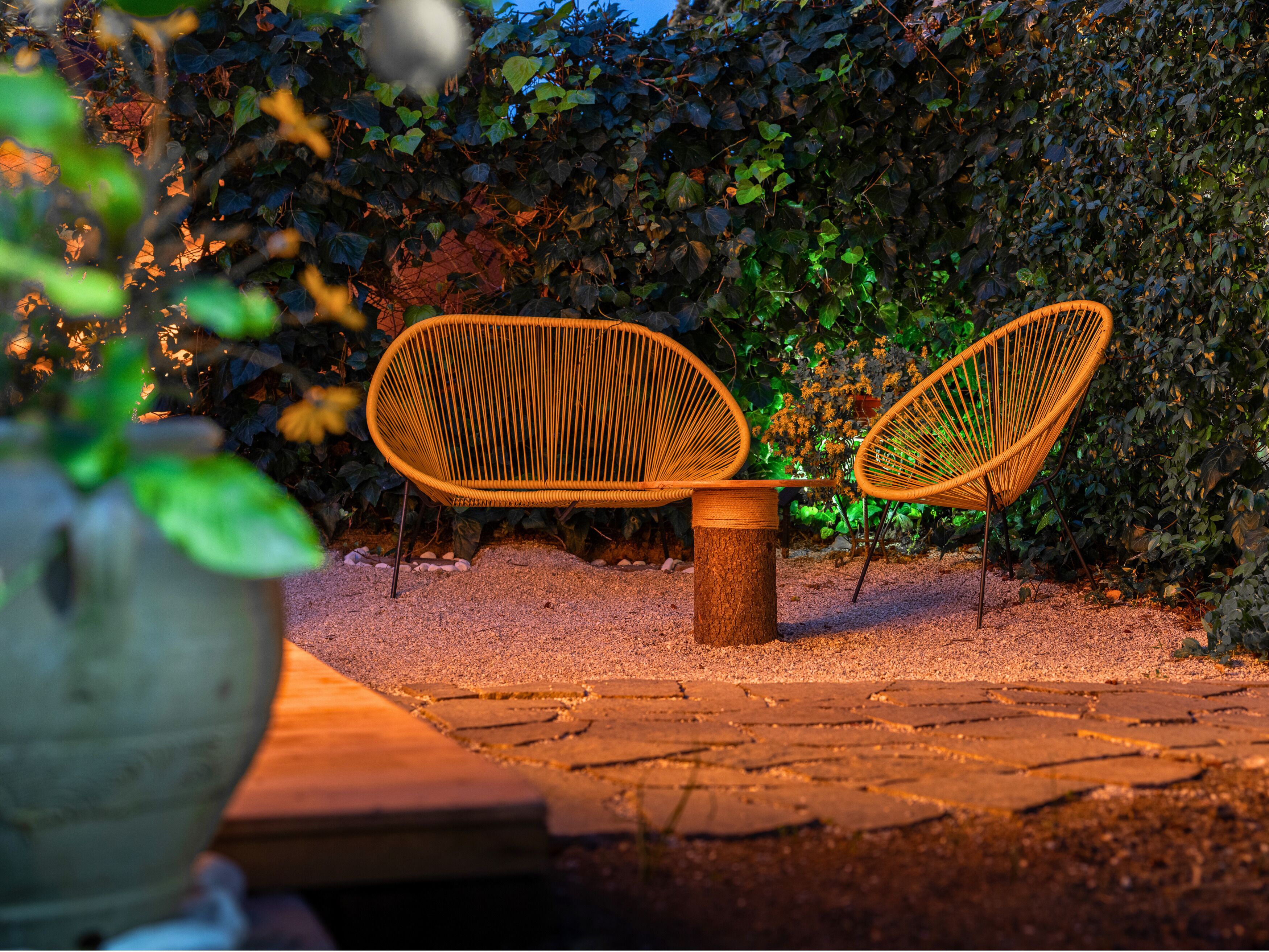 Two wicker chairs with a small table, set on a gravel path with a hedge in the background.