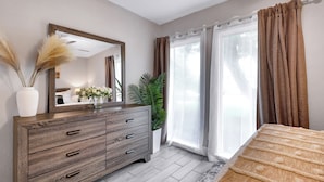 A bedroom with a wooden dresser, a mirror, a potted plant, and a window with curtains.