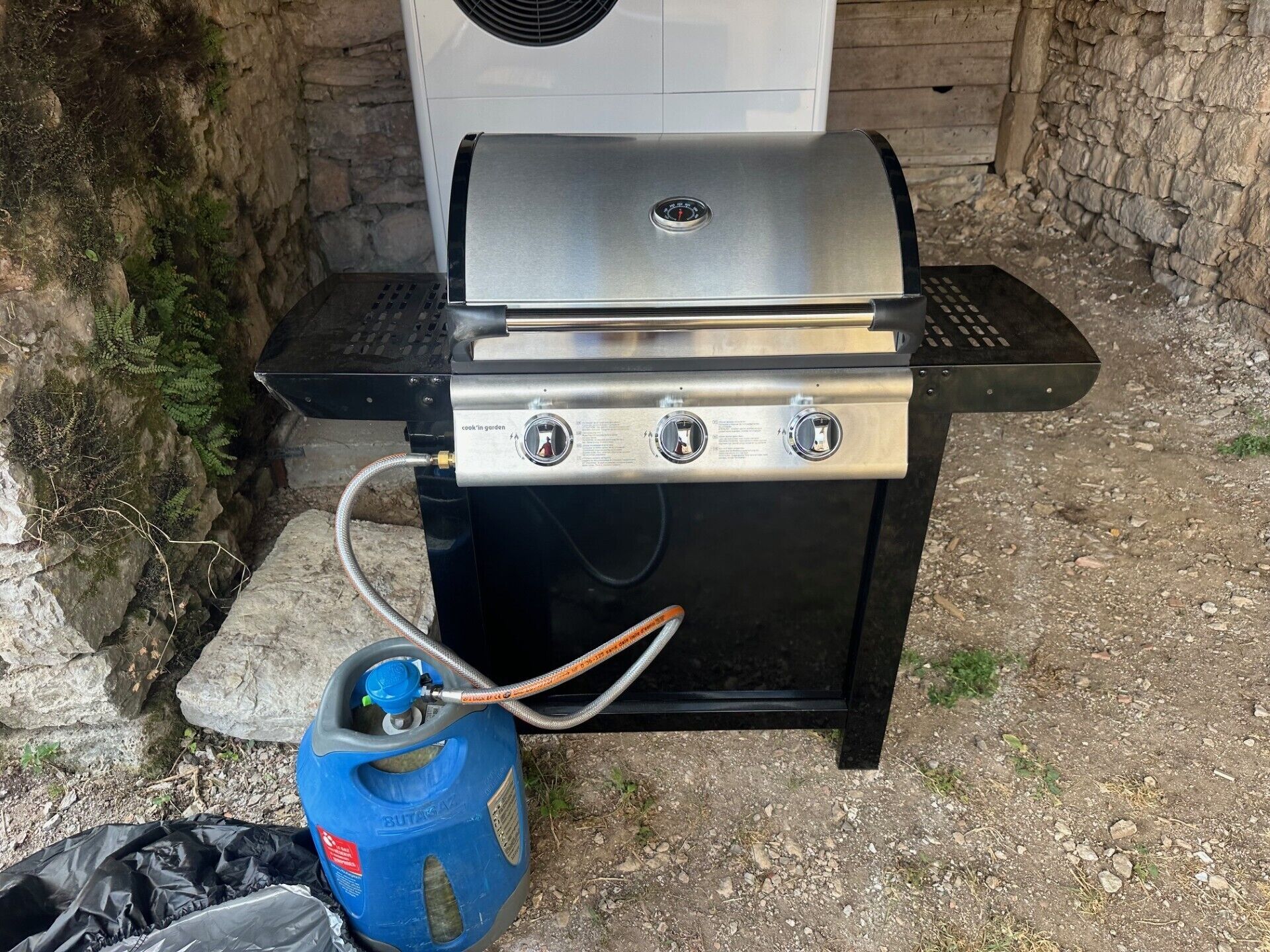 An outdoor grill with a gas tank, connected to a hose, and a vent fan.