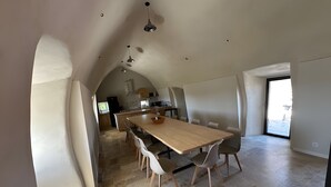 Un vaste espace de salle à manger avec une longue table et des chaises, une cuisine à l’arrière-plan et une grande fenêtre.