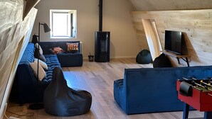Une chambre mansardée avec un canapé bleu, un fauteuil en forme de poire noir, un téléviseur posé sur un meuble et une cheminée.
