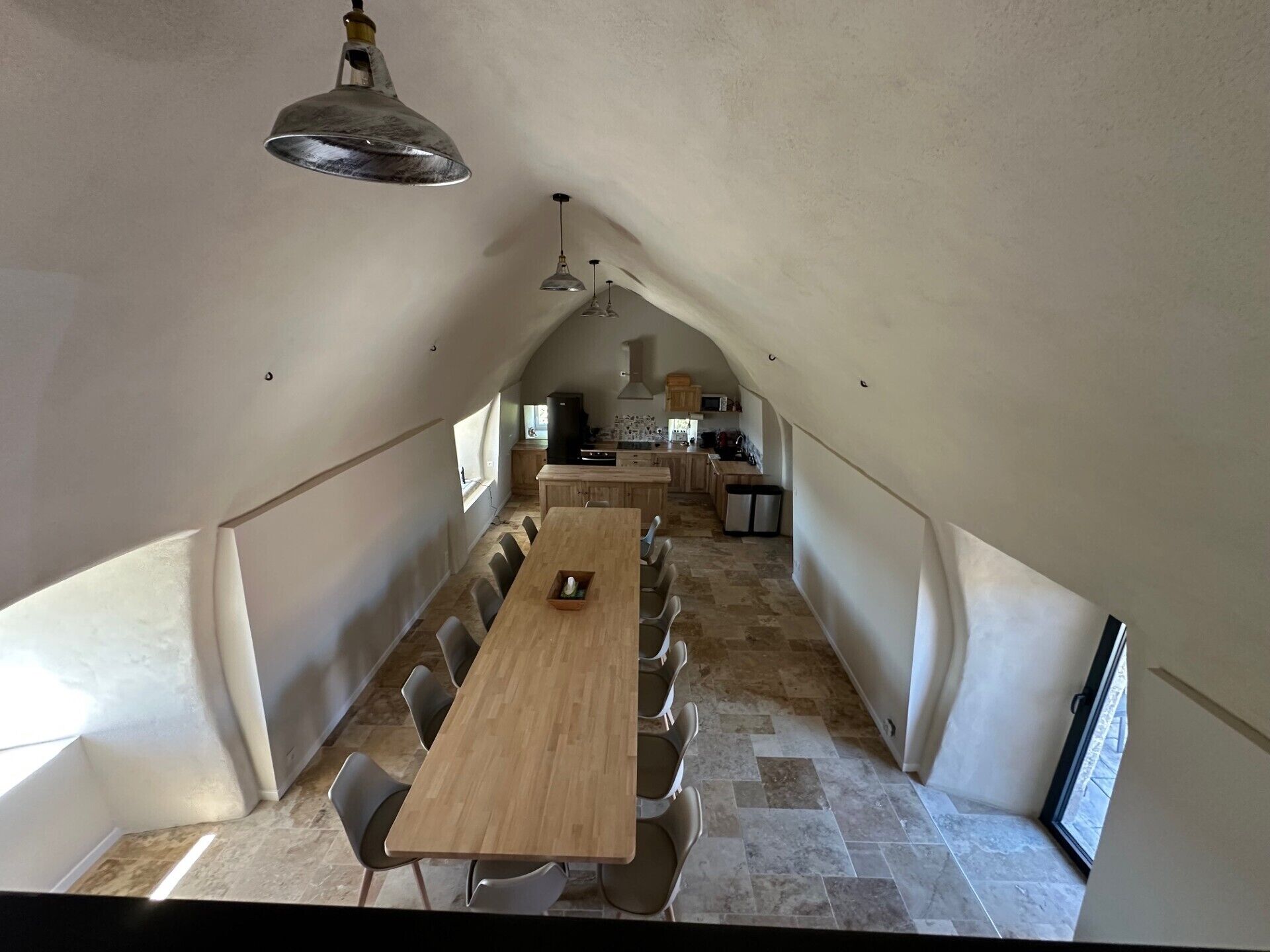 Un comedor con una larga mesa de madera y sillas, una cocina al fondo y un techo inclinado.