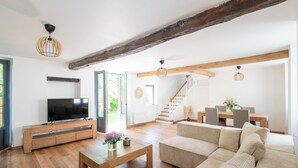 Un salon spacieux avec un meuble TV en bois, un grand canapé beige et une table basse en bois. Un escalier mène à un autre niveau, et on y trouve un coin repas avec une table et des chaises en bois.