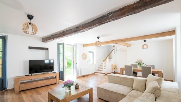 A spacious living room with a wooden TV stand, a large beige sofa, and a wooden coffee table. There is a staircase leading to another level, and a dining area with a wooden table and chairs. - Beautiful family house sleeps 9 near the castle in Dampierre (Dampierre-en-Yvelines)