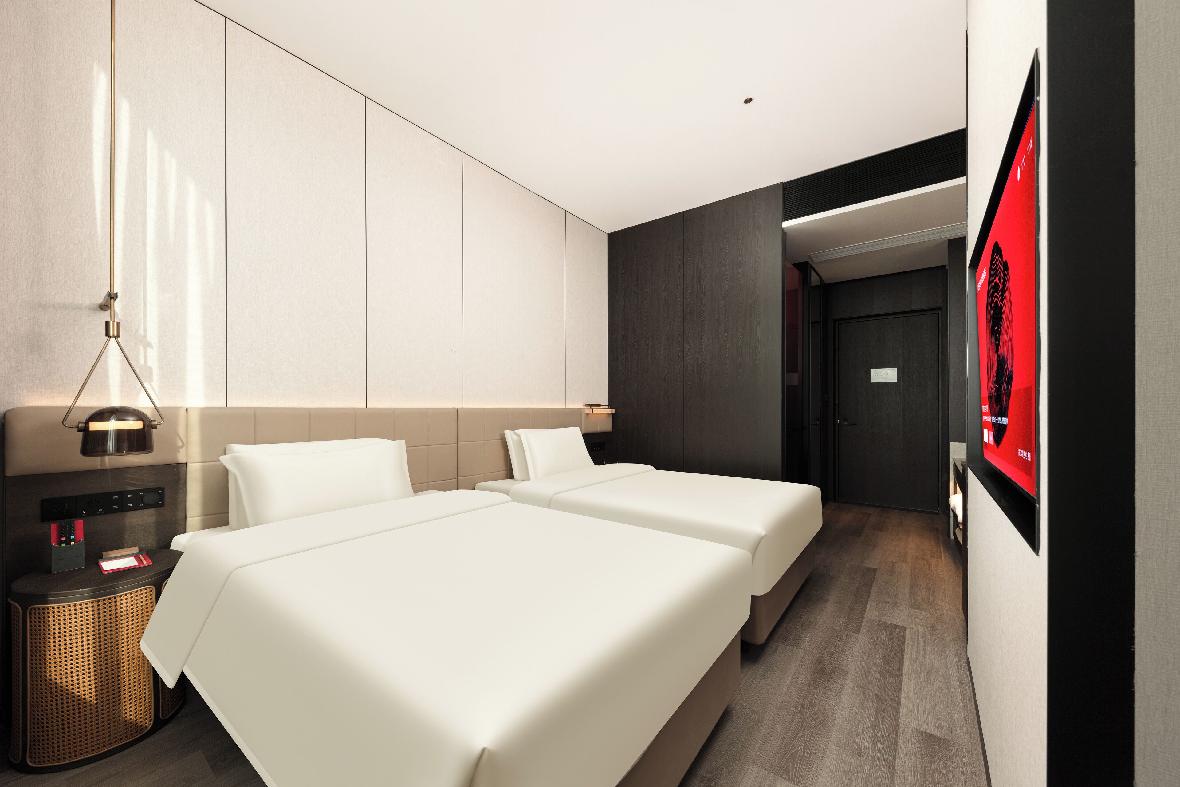 Une chambre d’hôtel avec deux lits, une table de chevet avec une lampe, un téléviseur fixé au mur et une porte donnant sur une autre pièce.