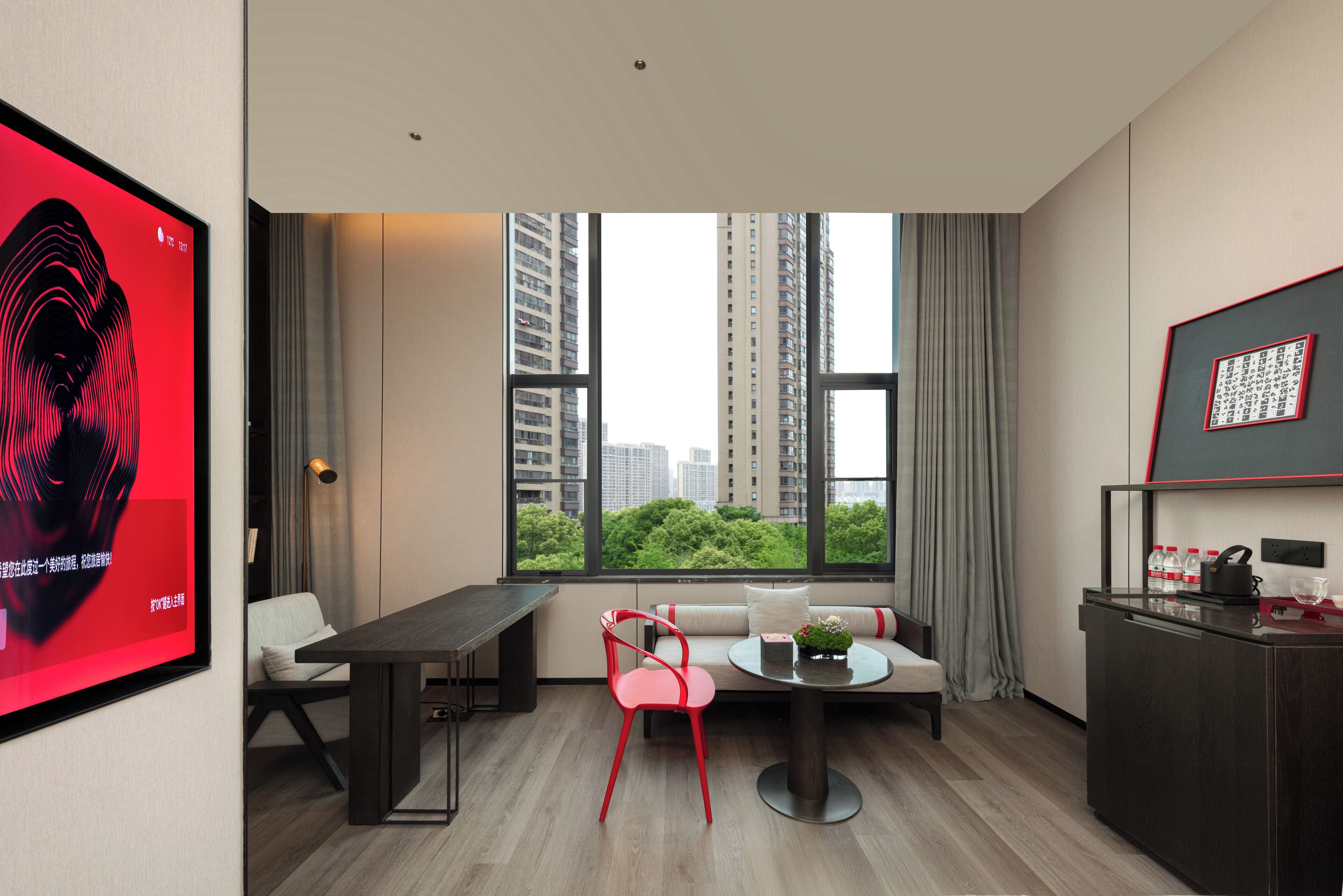 Een moderne hotelkamer met een zithoek, een bank, een rode stoel, een tafeltje met een plant, en uitzicht op hoge gebouwen en groen.