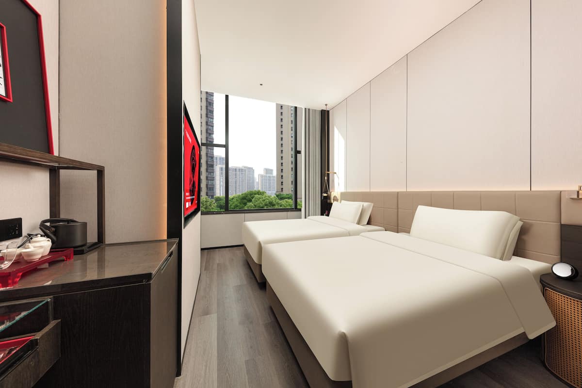 A modern hotel room with two beds, a desk, and a large window offering a city view.