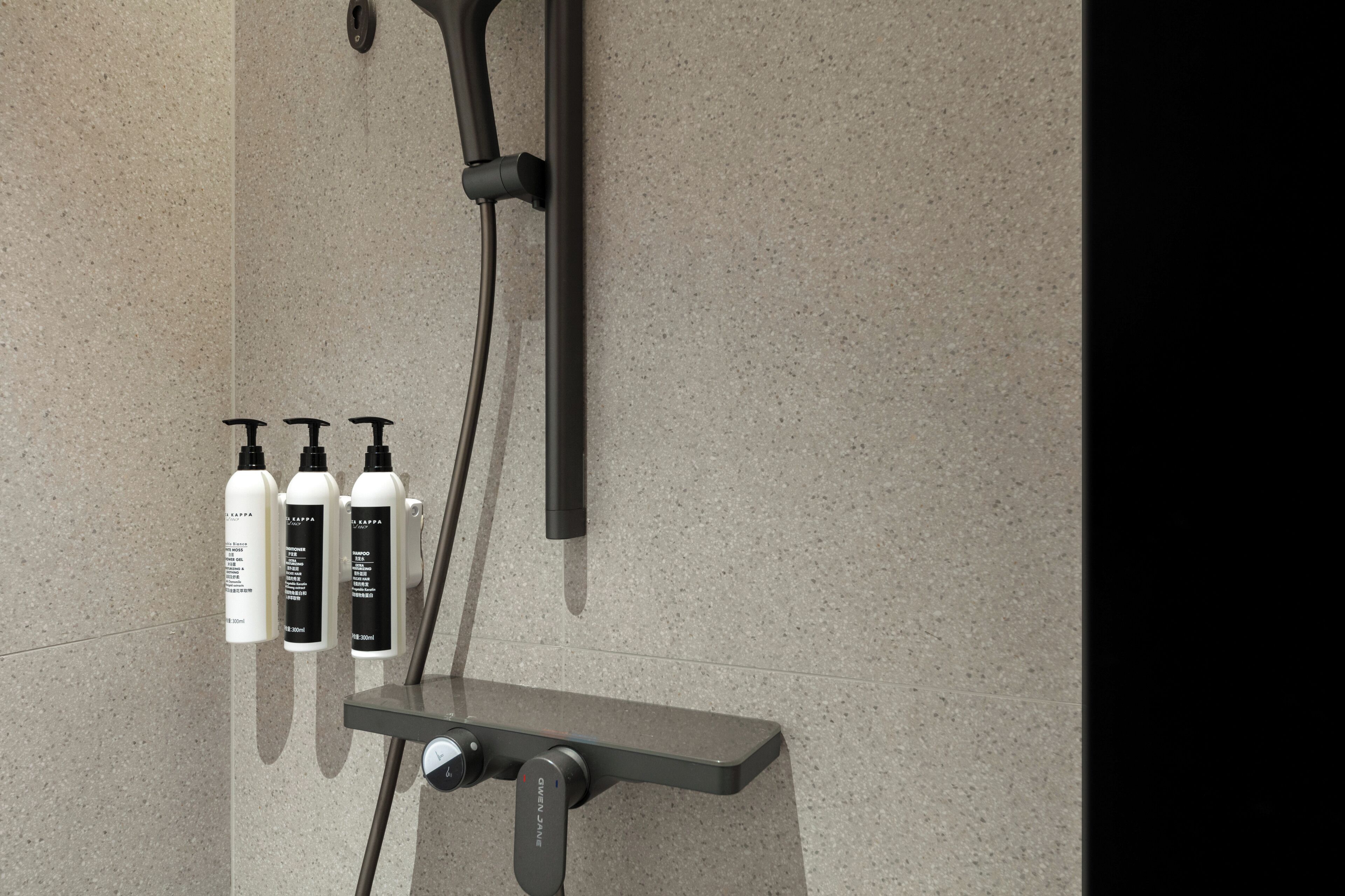 A shower with a handheld showerhead and a wall-mounted one, a shelf with three bottles of shampoo, and a control knob.