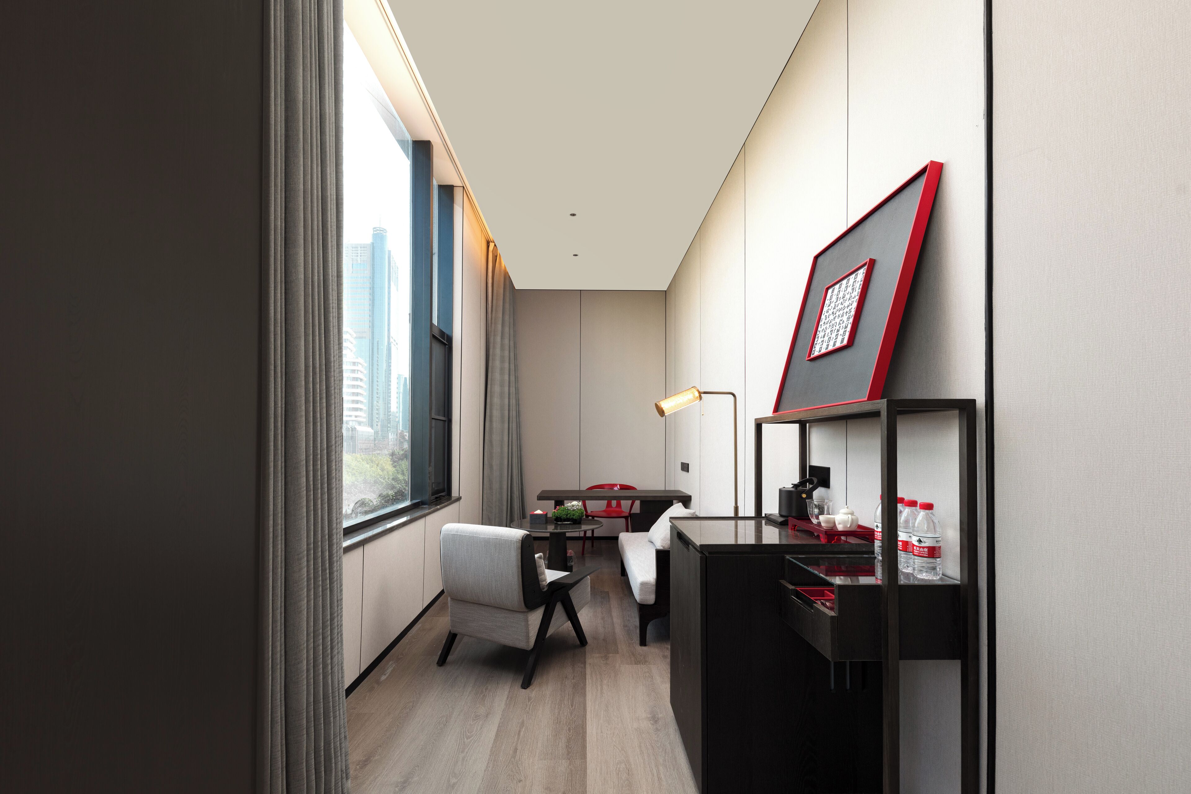 A modern hotel room with a desk, chair, and a small table with a lamp. There is a window with a view of buildings outside.