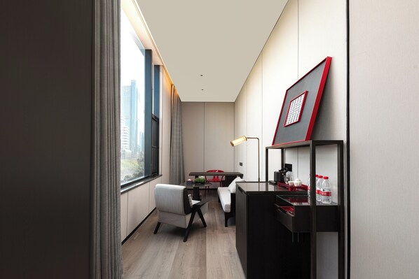 A modern hotel room with a desk, chair, and a small table with a lamp. There is a window with a view of buildings outside.