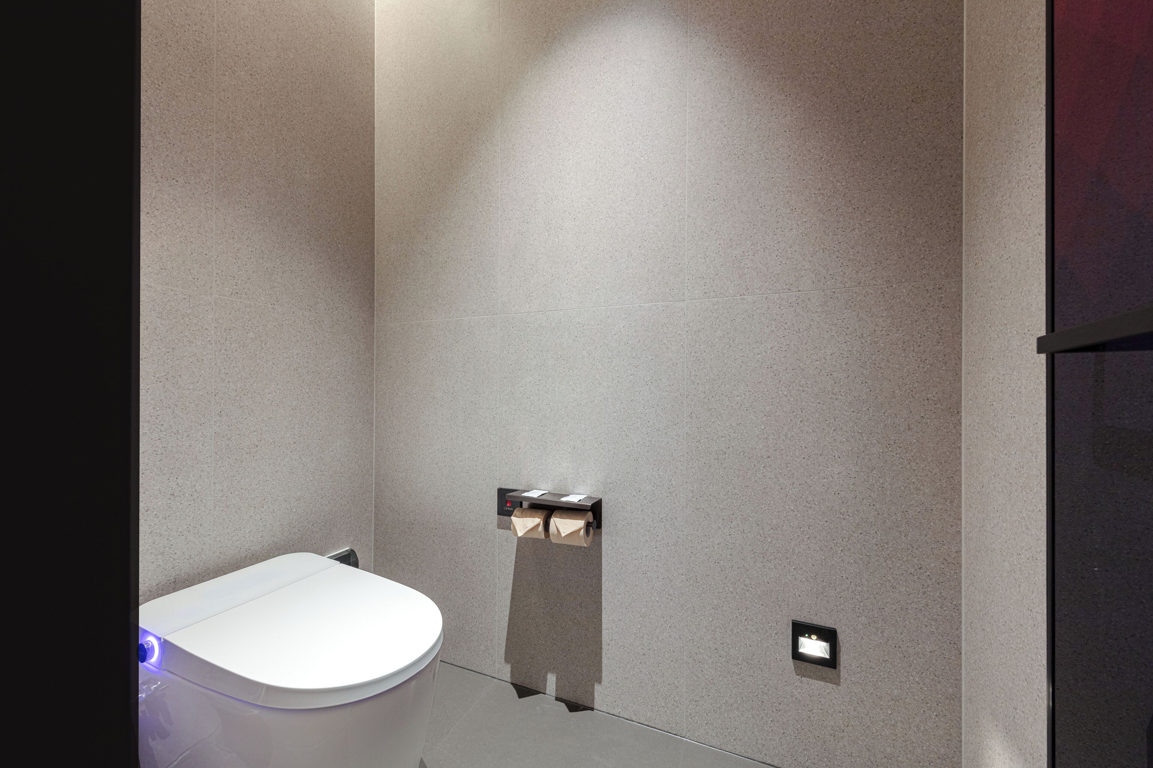 A modern bathroom with a white toilet, a towel dispenser, and a wall-mounted light fixture.