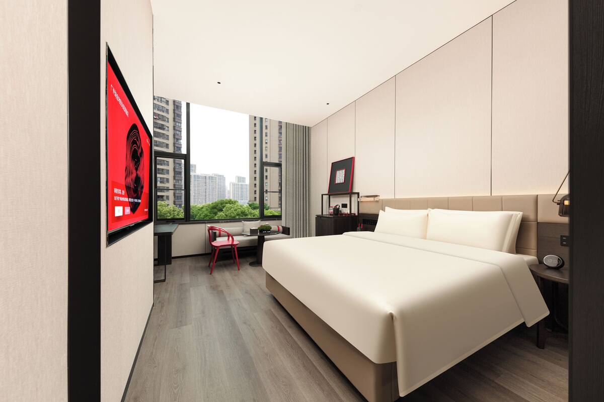 A modern hotel room with a large bed, a desk, and a chair. There is a window with a view of buildings and greenery.