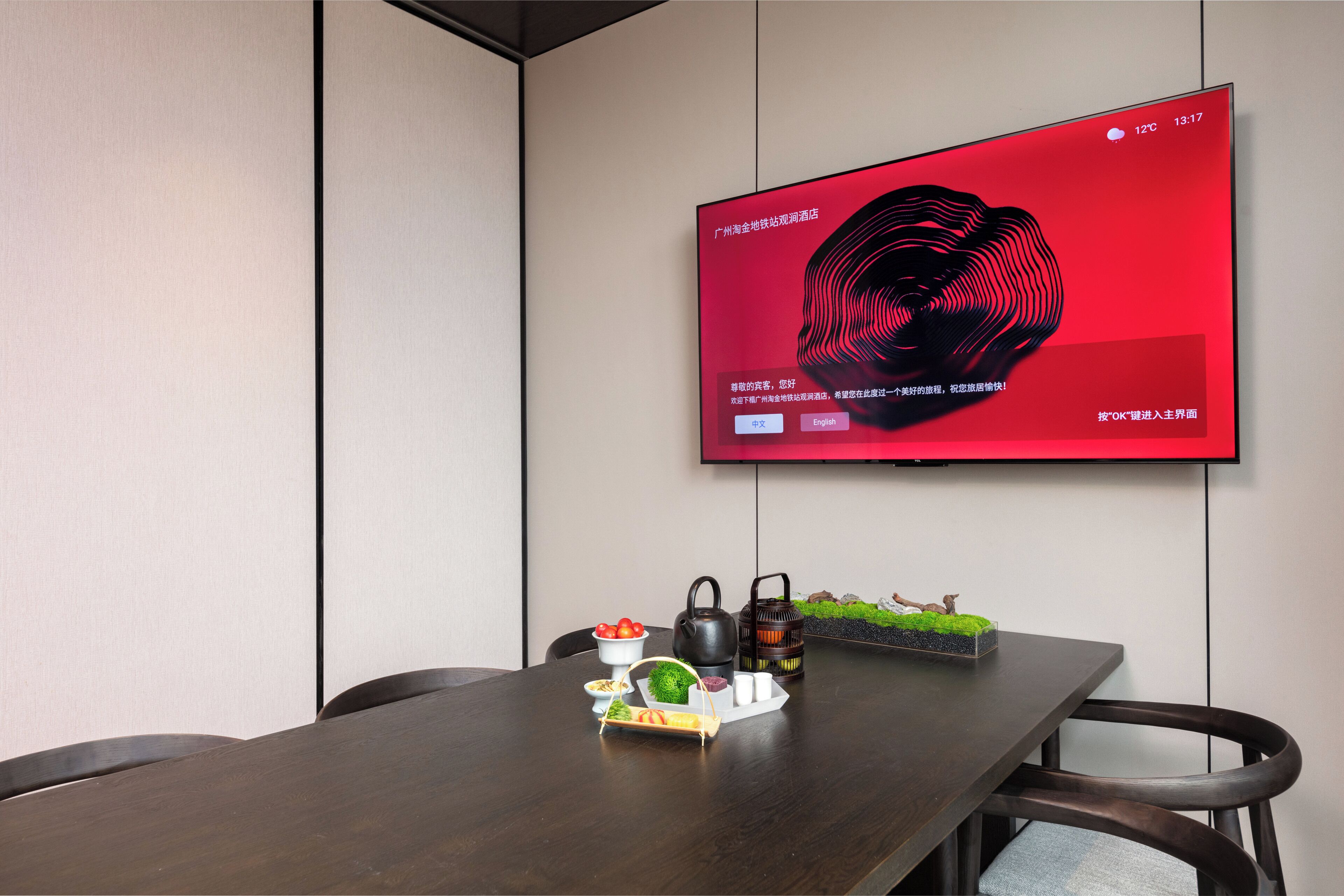 Un salón de conferencias con una gran pantalla plana montada en la pared mostrando un gráfico rojo. En el centro hay una mesa de madera con un pequeño arreglo decorativo.