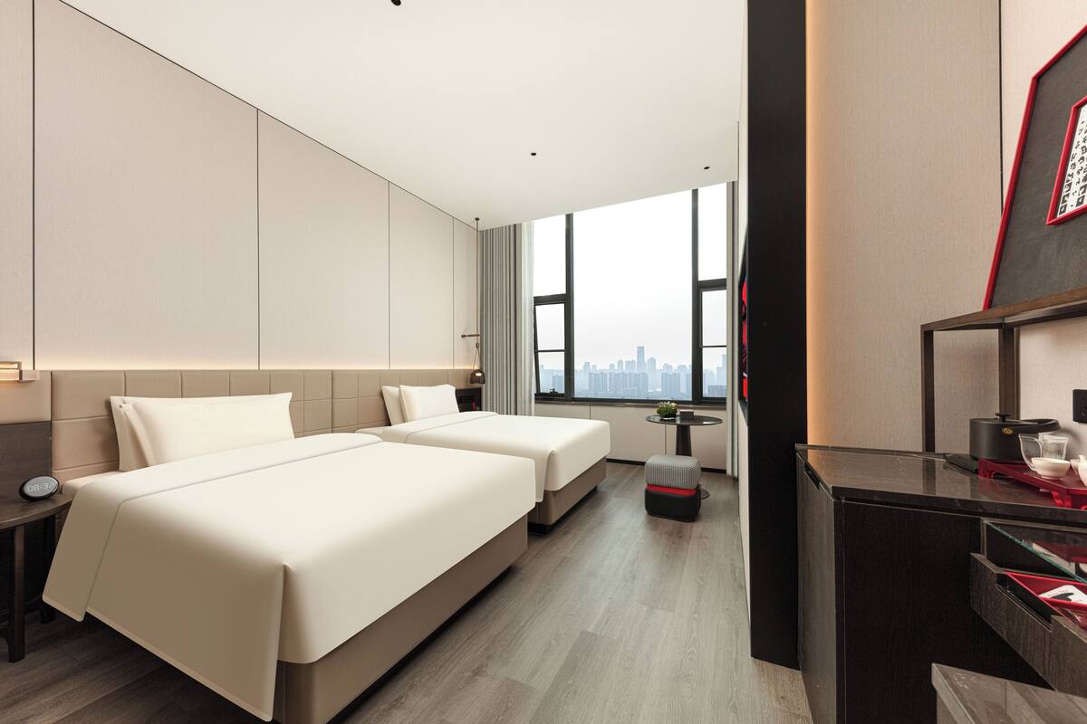 A modern hotel room with two beds, a desk, and a view of the city skyline.