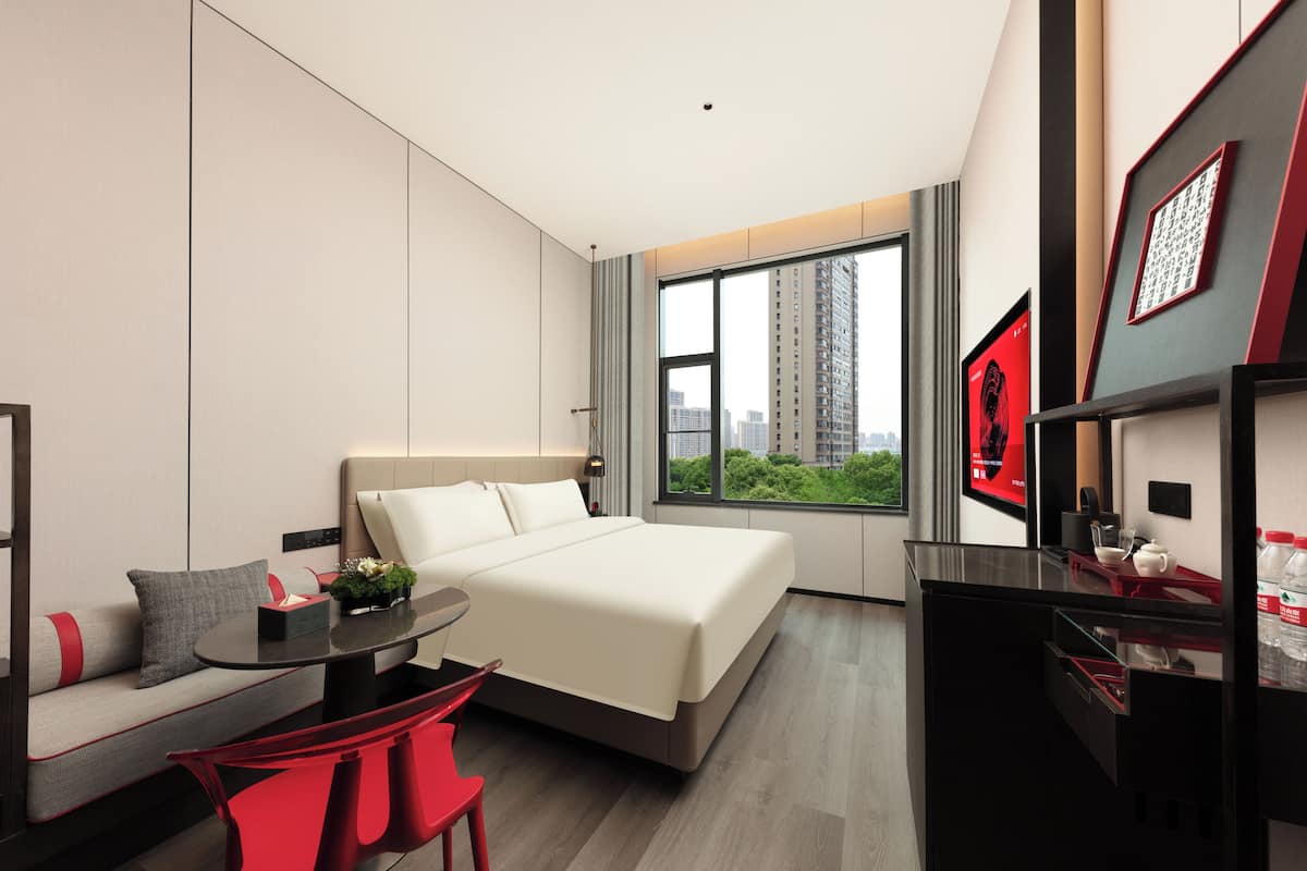 A modern hotel room with a bed, a desk, a chair, and a large window offering a city view.