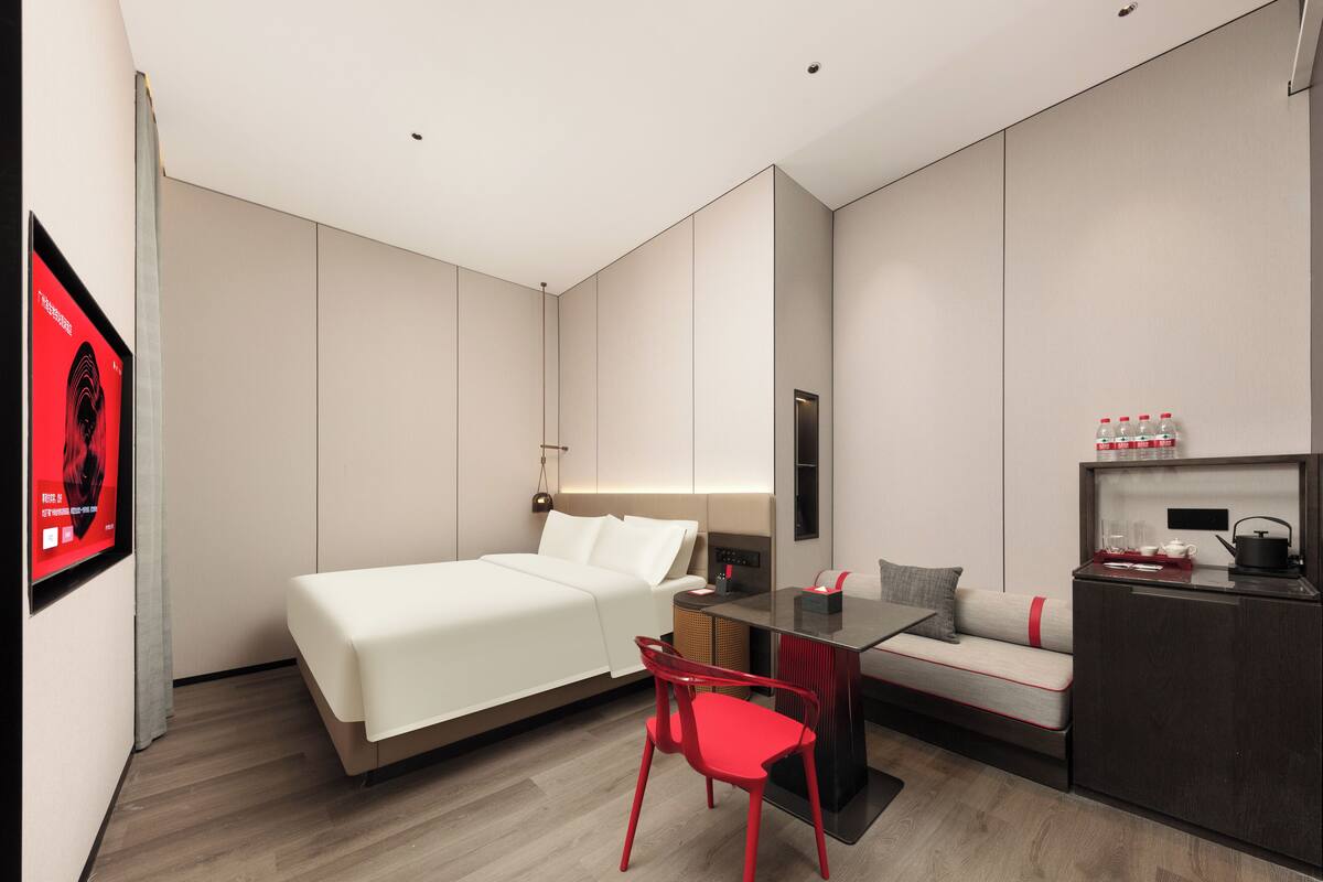 A modern hotel room with a bed, a desk, a red chair, a sofa, and a minibar.