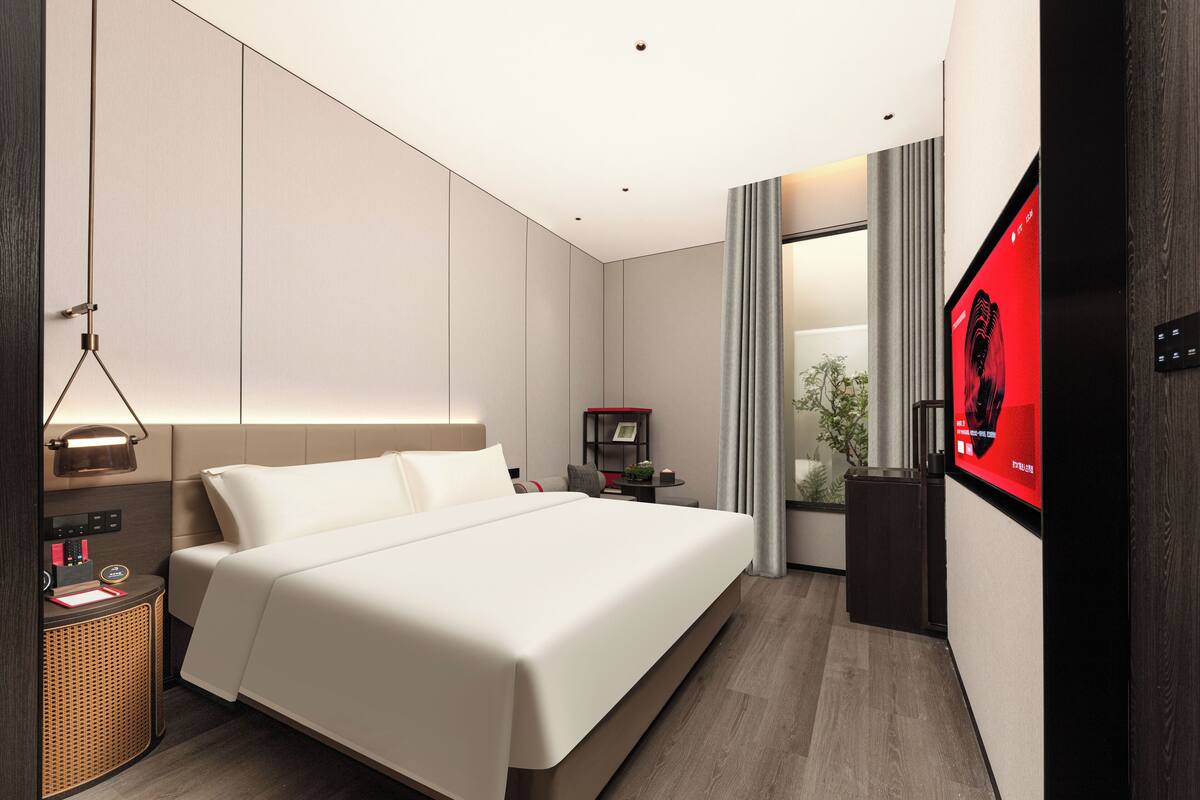 A modern hotel room with a large bed, a bedside table with a lamp, a TV, and a small desk with a chair.