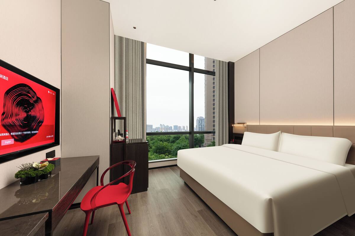 A modern hotel room with a large bed, a desk with a chair, a TV, and a view of the city.