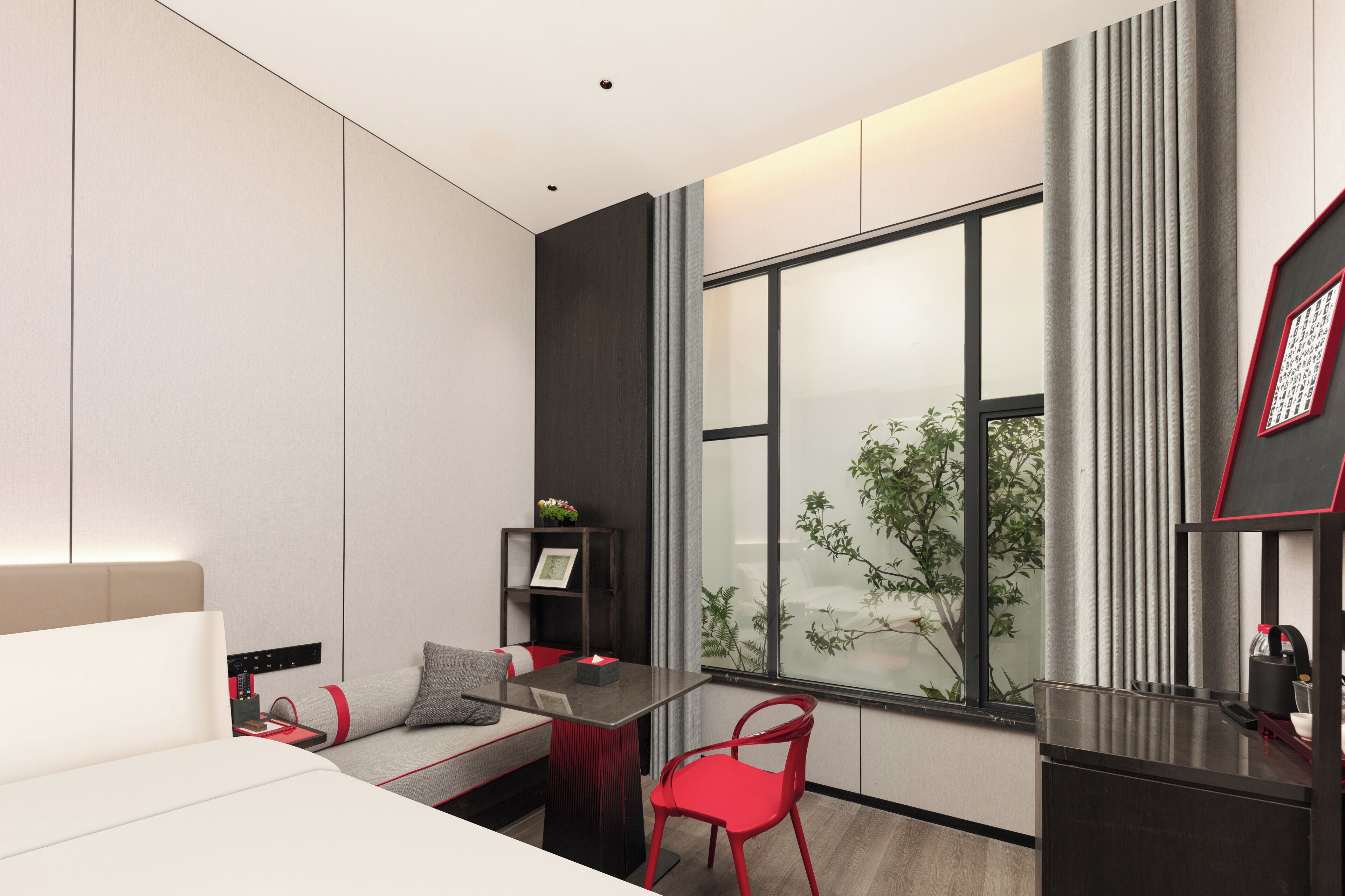 A modern hotel room with a bed, a small table, a red chair, a desk, and a large window with a view of greenery outside.