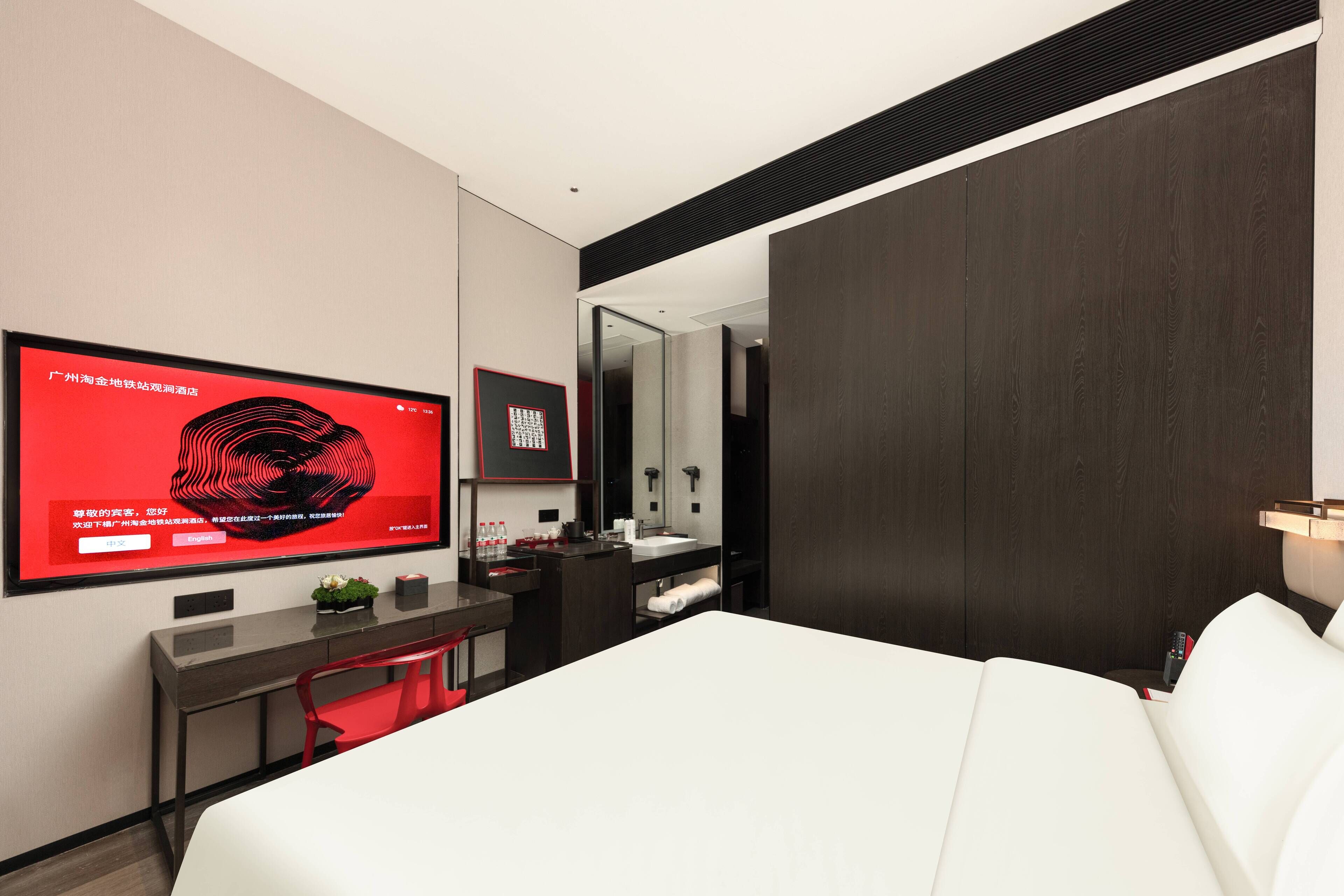 Una habitación de hotel moderna con una cama grande, un escritorio con computadora, televisor y baño con espejo y lavamanos.