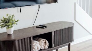 Un soggiorno moderno con una grande TV a schermo piatto montata a parete, un tavolino consolle in legno scuro con una pianta in vaso, e una scala sullo sfondo.