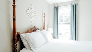 Un lit bien fait, avec une tête de lit en bois, un mur blanc agrémenté d’un élément décoratif et une fenêtre ornée de rideaux bleu clair.