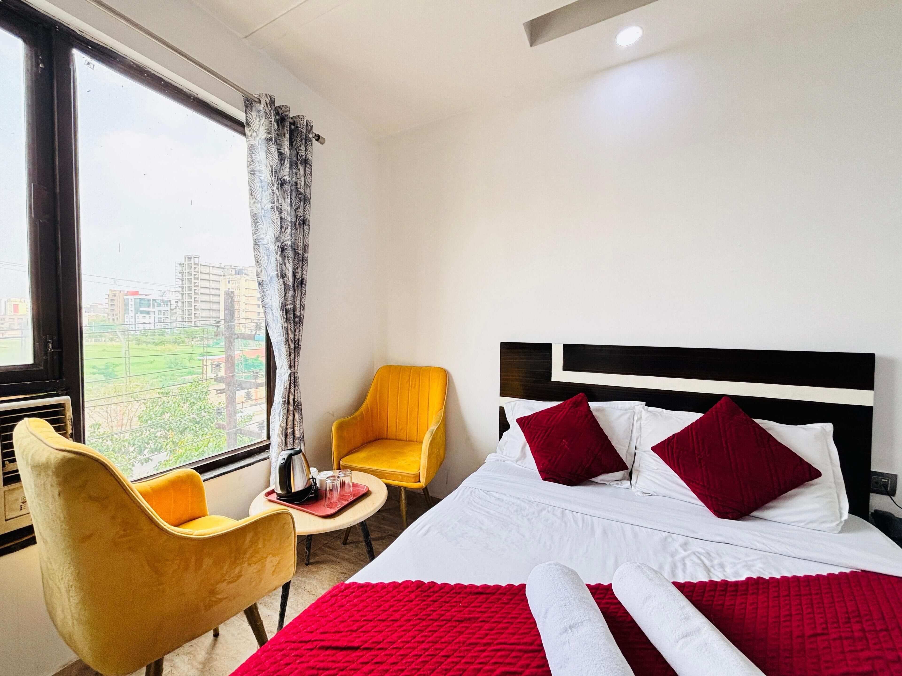A hotel room with a bed, two yellow chairs, a small table with a kettle, and a view of buildings through a window.