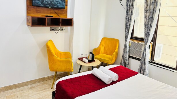 Une chambre d’hôtel avec un lit, deux chaises jaunes, une petite table, un téléviseur fixé sur un panneau en bois et une fenêtre avec des rideaux.