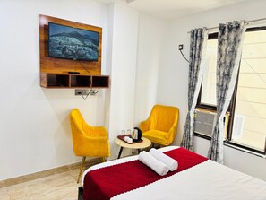 A hotel room with a bed, two yellow chairs, a small table, a TV mounted on a wooden panel, and a window with curtains. - Hotel Soho 62 (Noida)