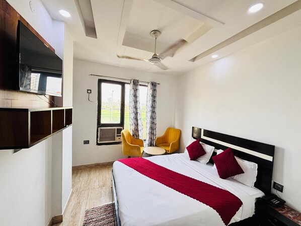 A hotel room with a bed, a TV mounted on the wall, a ceiling fan, a window with curtains, and a chair. - Hotel Soho 62 (Noida)