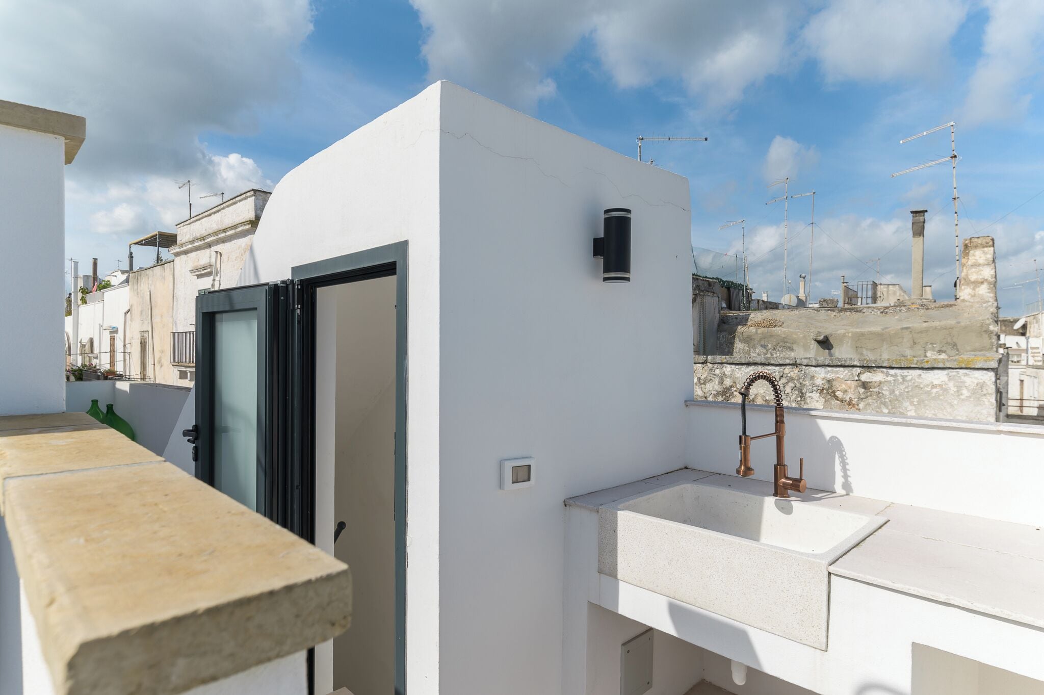 A rooftop terrace with a white wall, a black-framed door, a sink, and a small window.