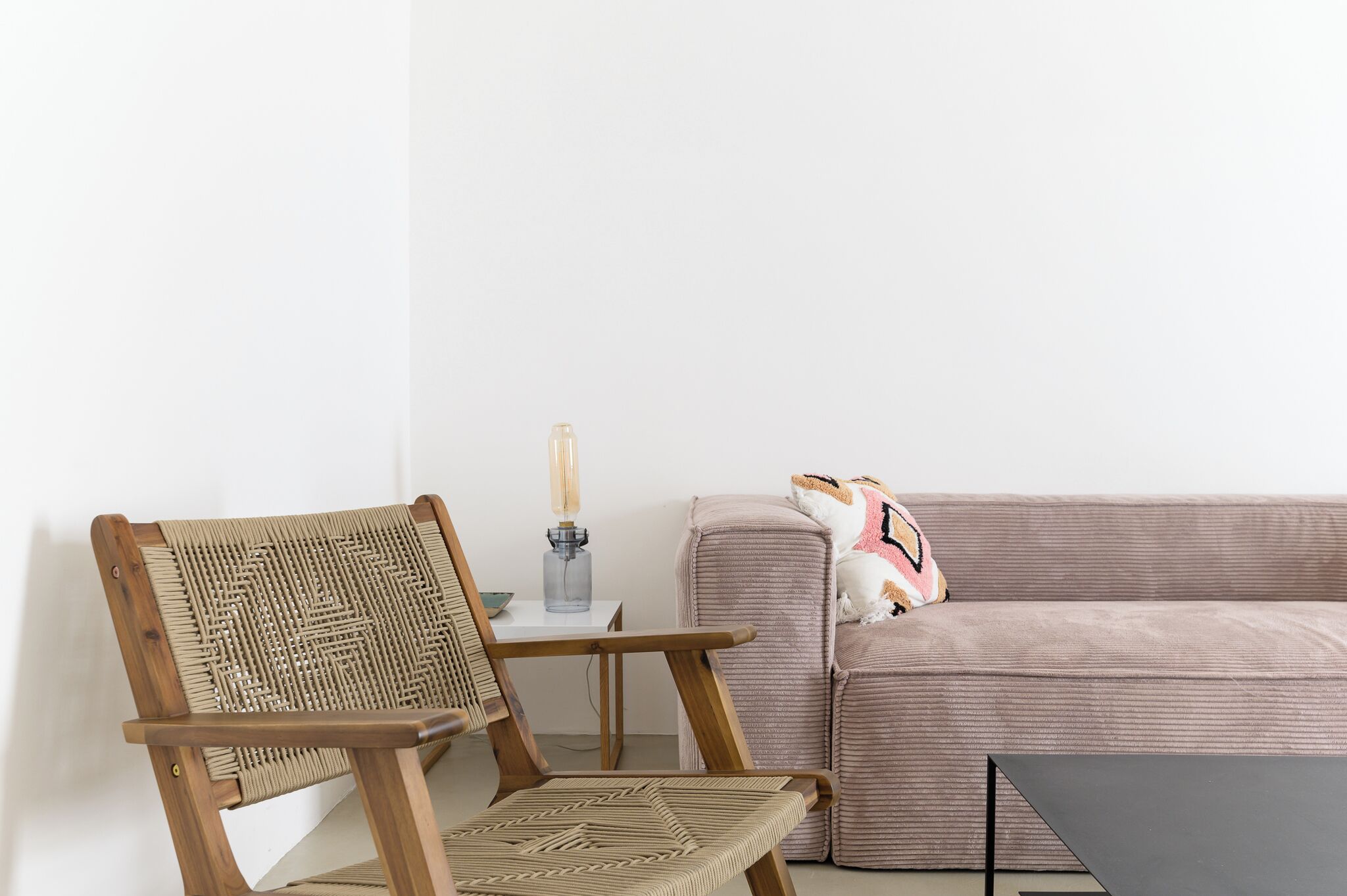 A wooden chair with a woven backrest, a small table with a glass bottle, and a sofa with a patterned cushion.
