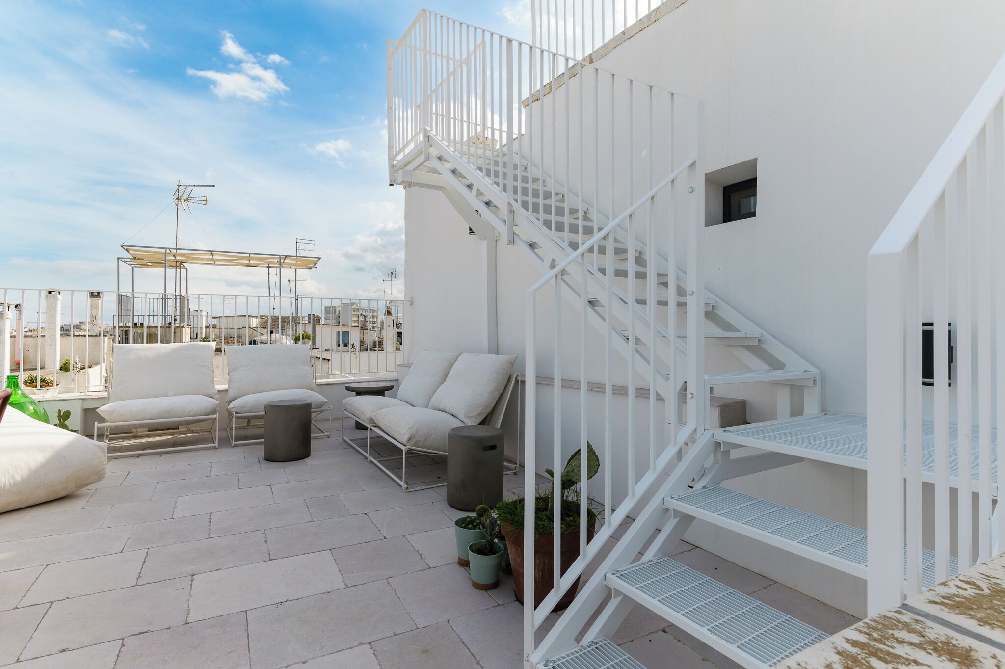 A rooftop terrace with white railings, a staircase, outdoor seating, and potted plants.