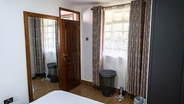Una habitación con cama, puerta de madera, ventana con cortinas y un basurero.