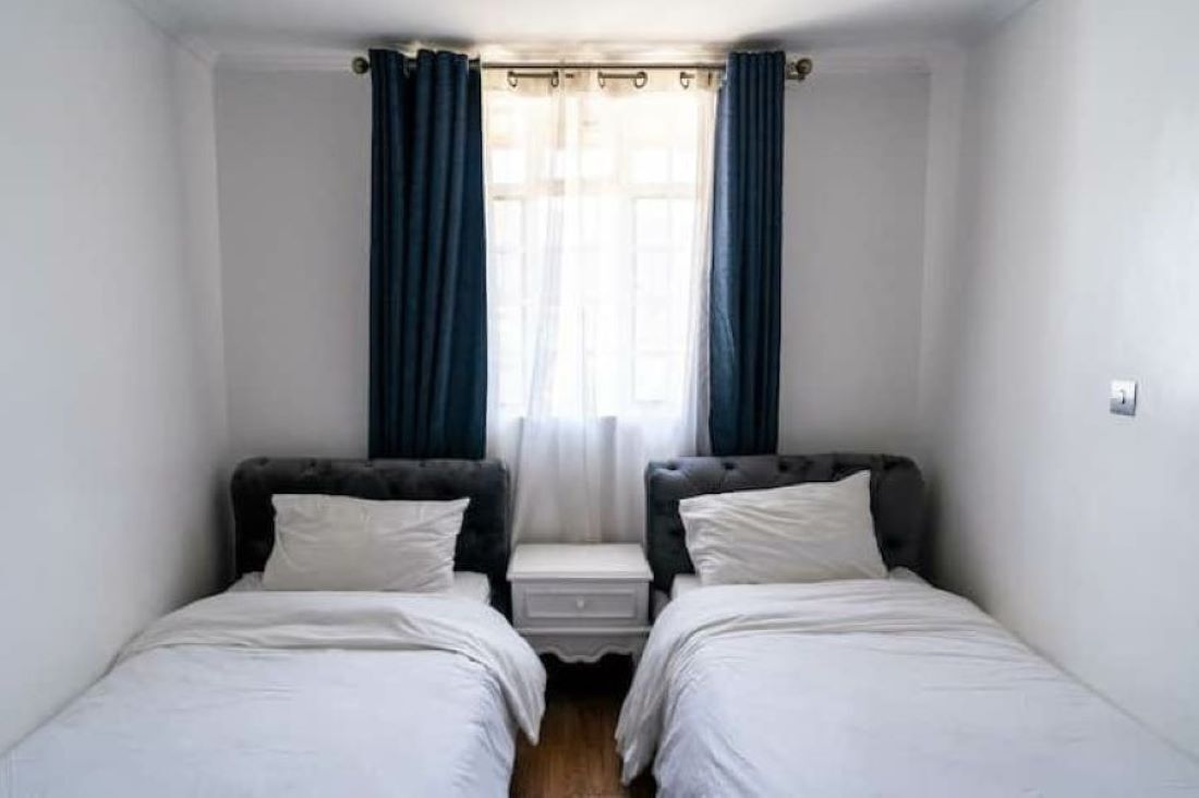 A small, clean room with two single beds, a nightstand, and a window with curtains.