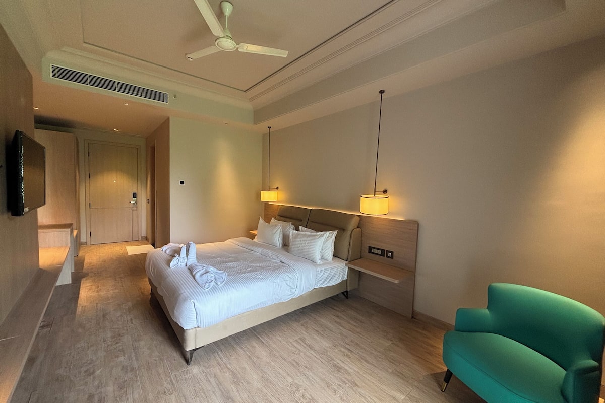A hotel room with a bed, a green armchair, a ceiling fan, and a TV on the wall.