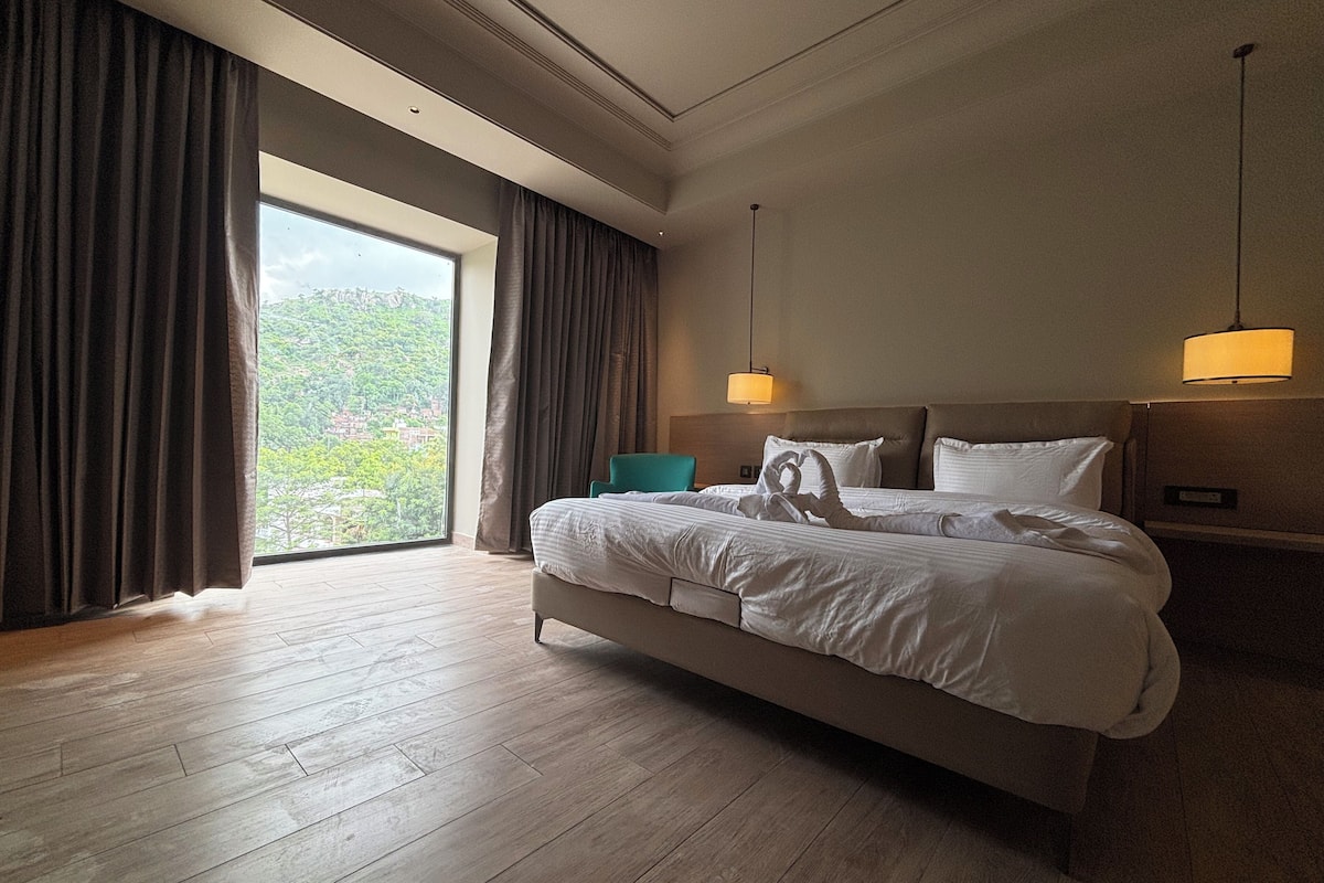 A hotel room with a large bed, a view of greenery outside, and a modern design.