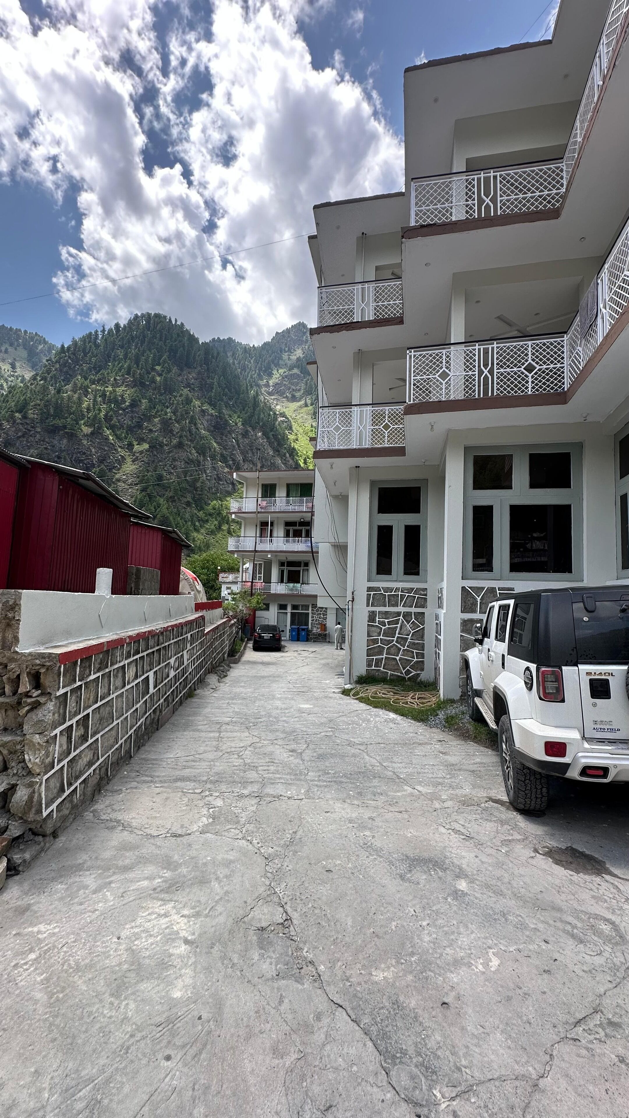 A multi-story building with balconies, a parking area with a white SUV, and a mountainous backdrop.