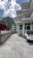 Un edificio de varios pisos con balcones, un estacionamiento con un SUV blanco y montañas al fondo.