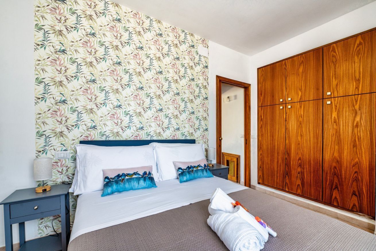 Un dormitorio con papel tapiz floral, un armario grande de madera, una cama bien hecha con almohadas y una mesita de noche con una lámpara.