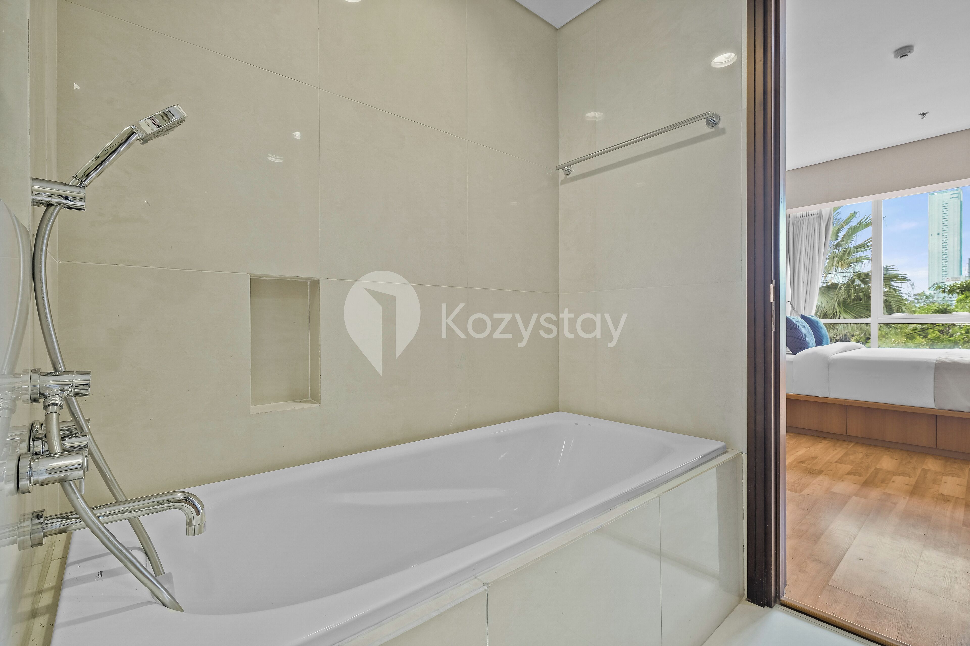 A modern bathroom with a large white bathtub, chrome fixtures, and a view of a balcony with a wooden floor and greenery.