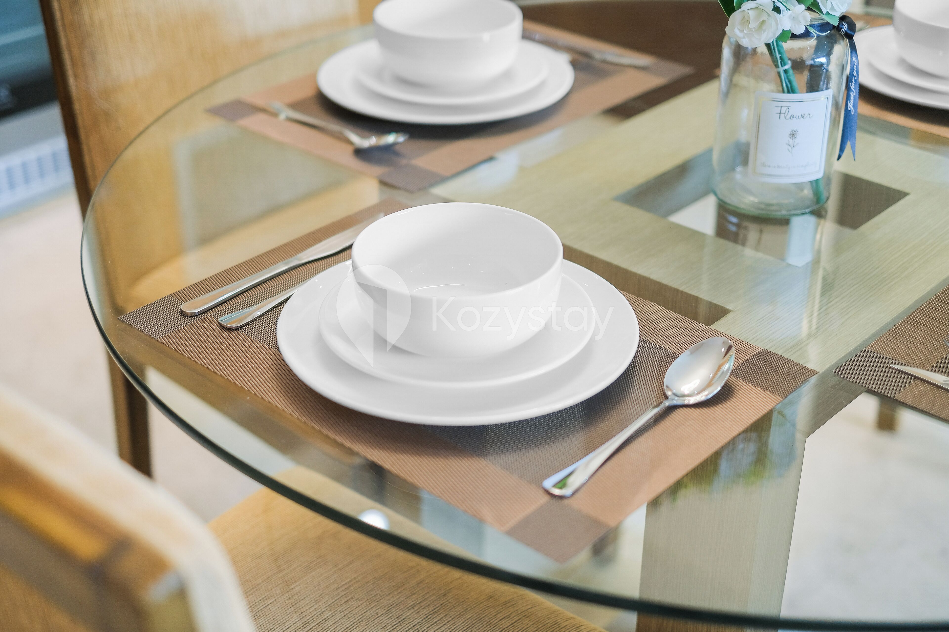 Une table à manger avec des assiettes blanches, un plateau en verre et un vase rempli de fleurs.