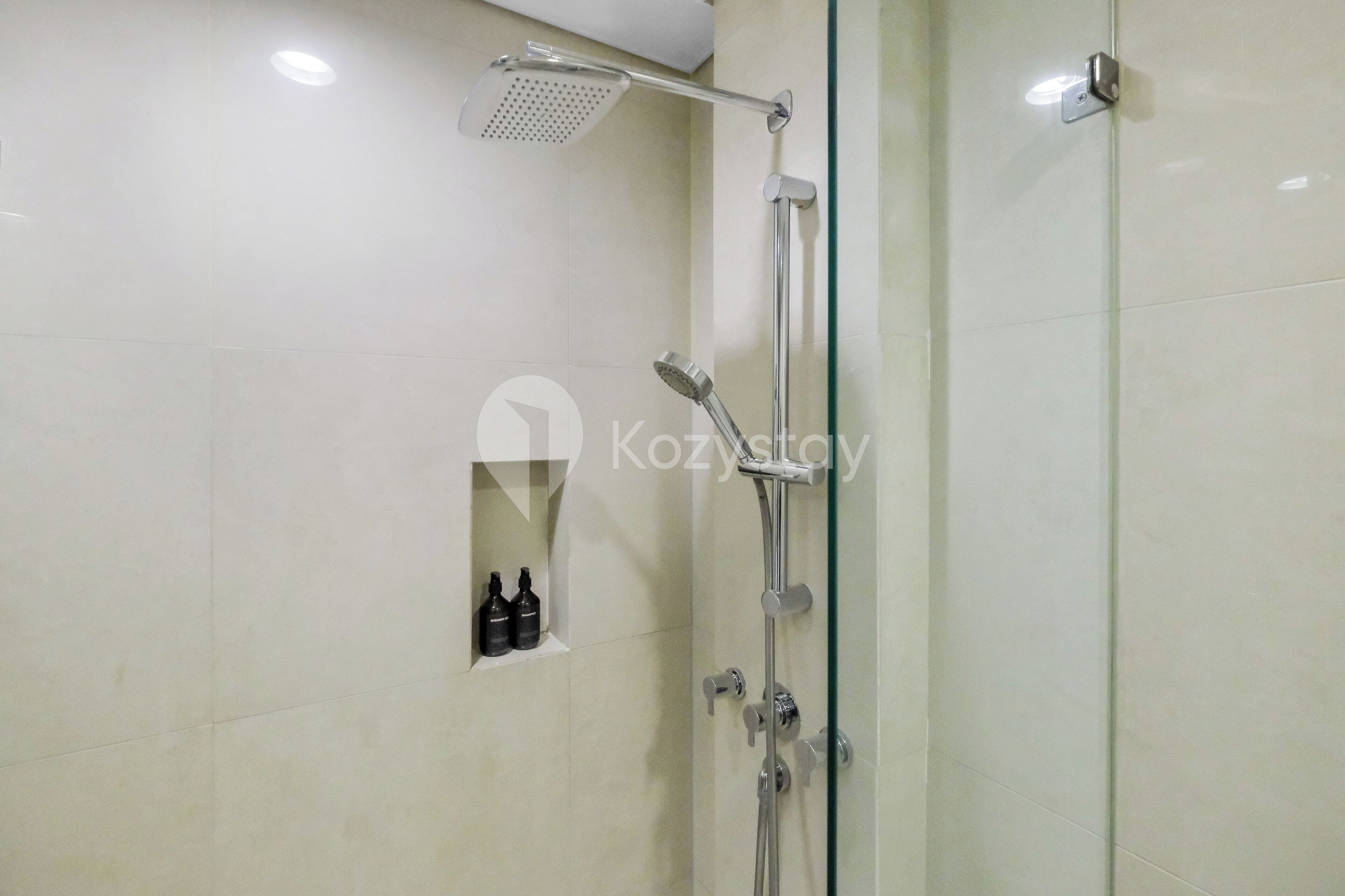 A modern shower area with a handheld showerhead, a fixed showerhead, and a glass-enclosed shower stall. There are two bottles of shampoo and conditioner on the wall.