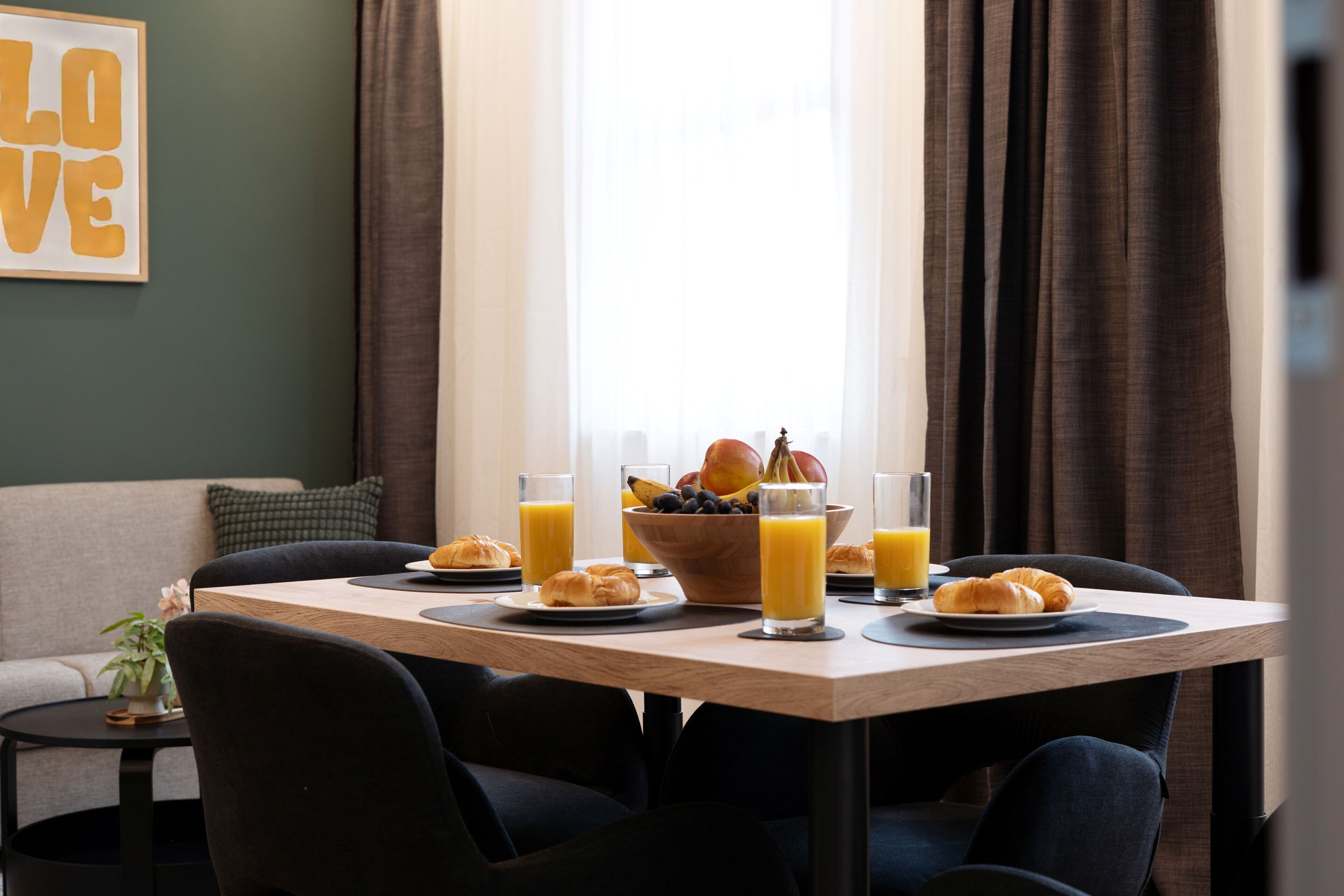 A breakfast setup with a wooden table, black chairs, a bowl of fruit, croissants, and glasses of orange juice.