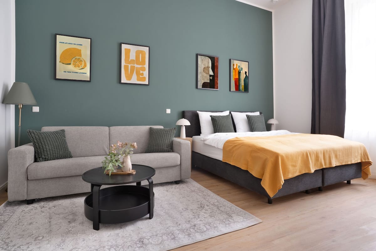 Une chambre moderne avec un canapé gris, un lit recouvert d’une couverture jaune et des œuvres d’art accrochées aux murs.