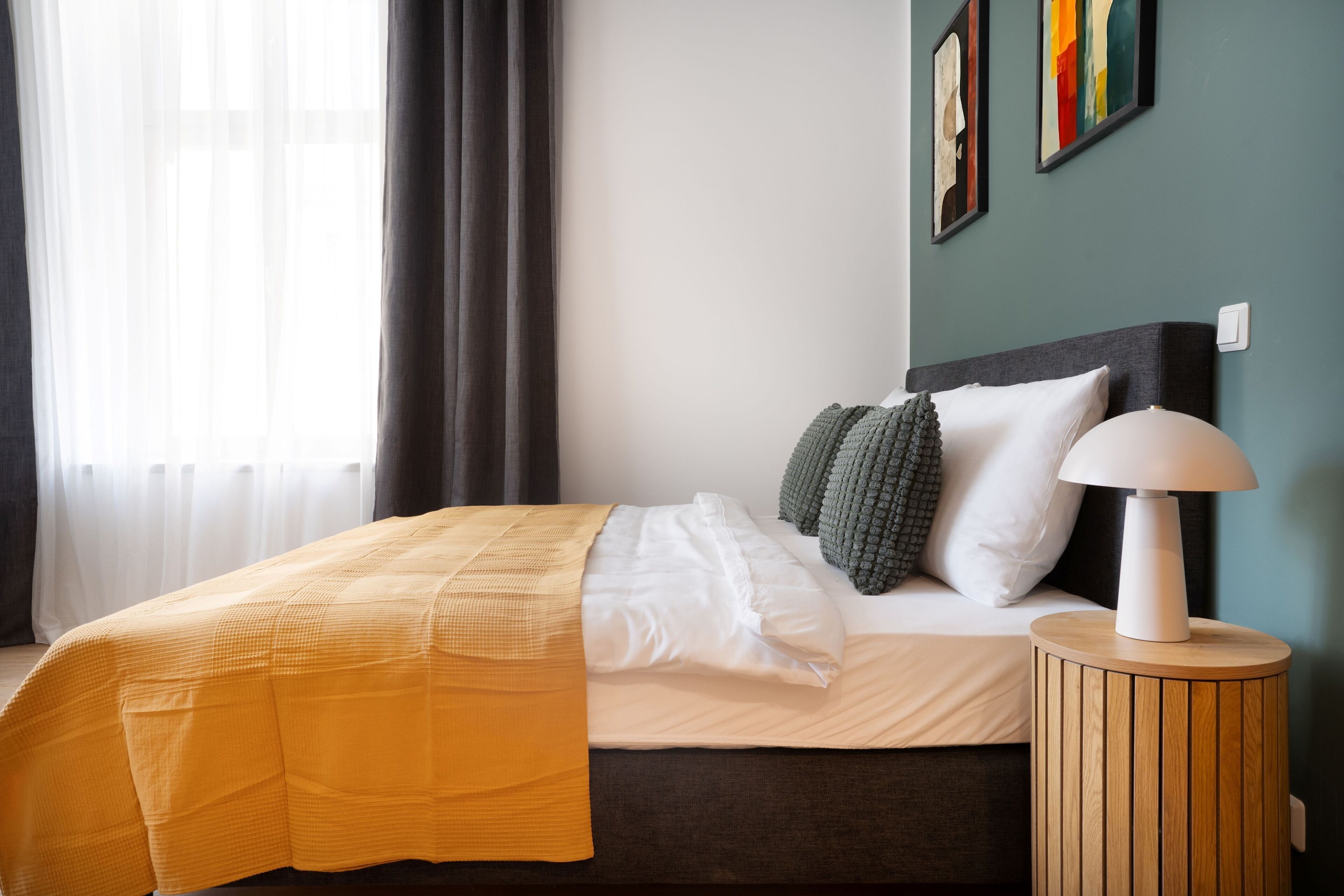 Un letto rifatto con una coperta gialla, un cuscino bianco e un cuscino grigio su una testiera scura. Accanto al letto, un comodino rotondo in legno con una lampada bianca.
