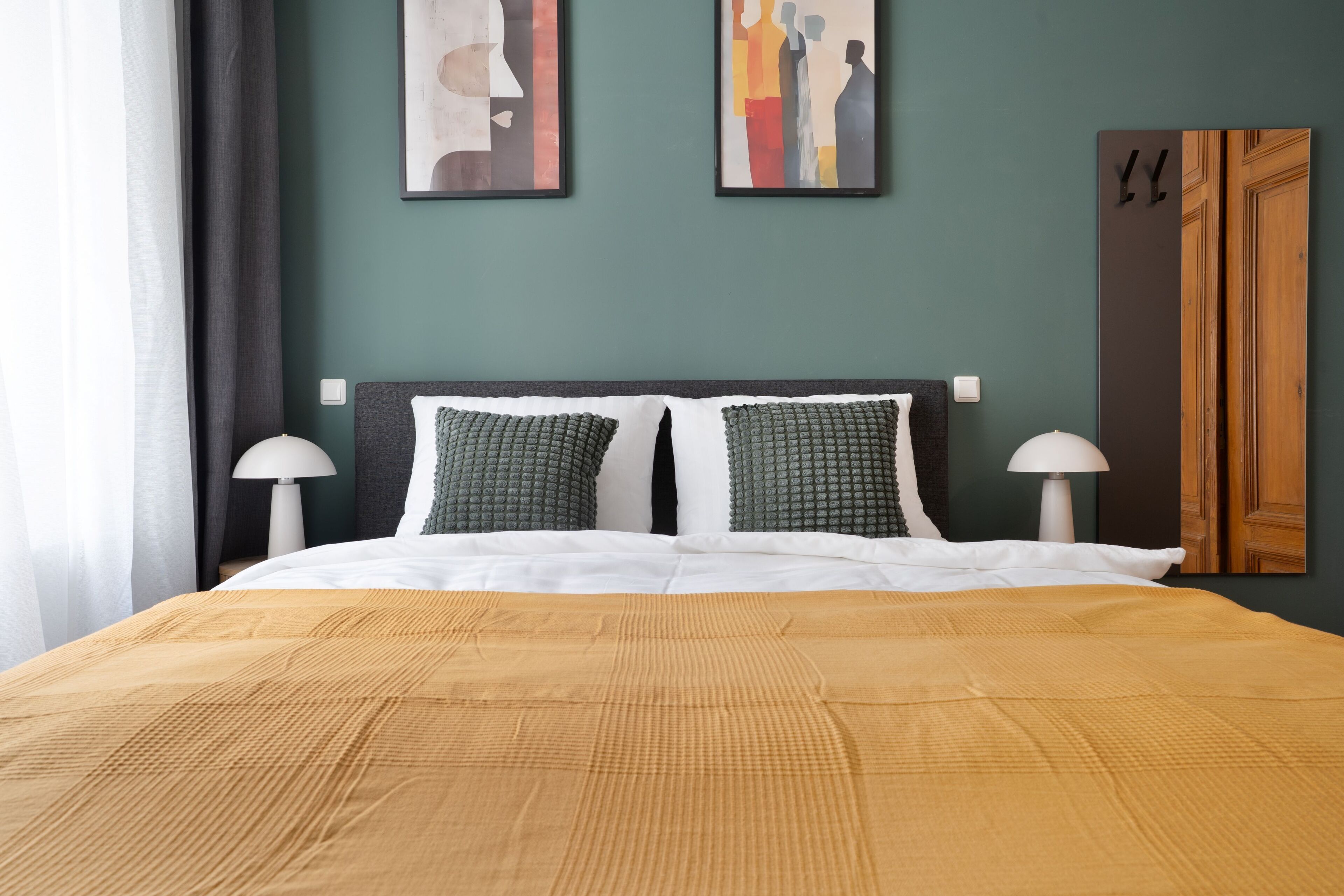 A neatly made bed with a textured brown bedspread, two pillows, and two bedside lamps. The room has a green wall and a wooden door.