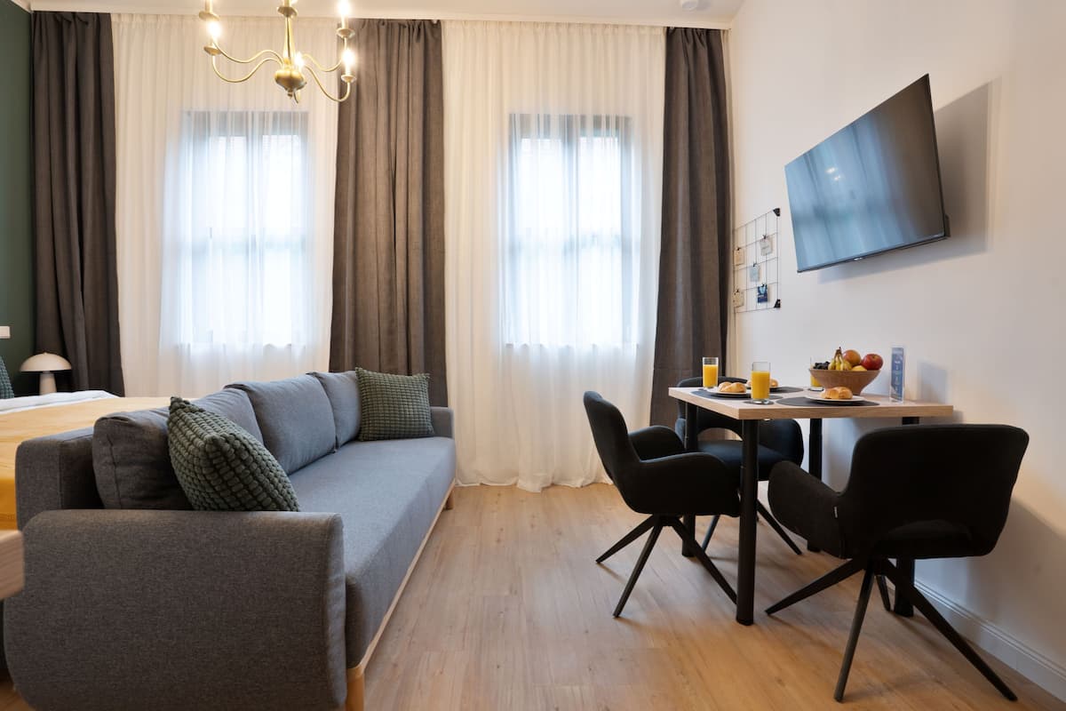 Un salon moderne avec un canapé gris, des chaises noires, une table à manger en bois sur laquelle sont disposés des fruits et des boissons, et un téléviseur à écran plat fixé au mur.