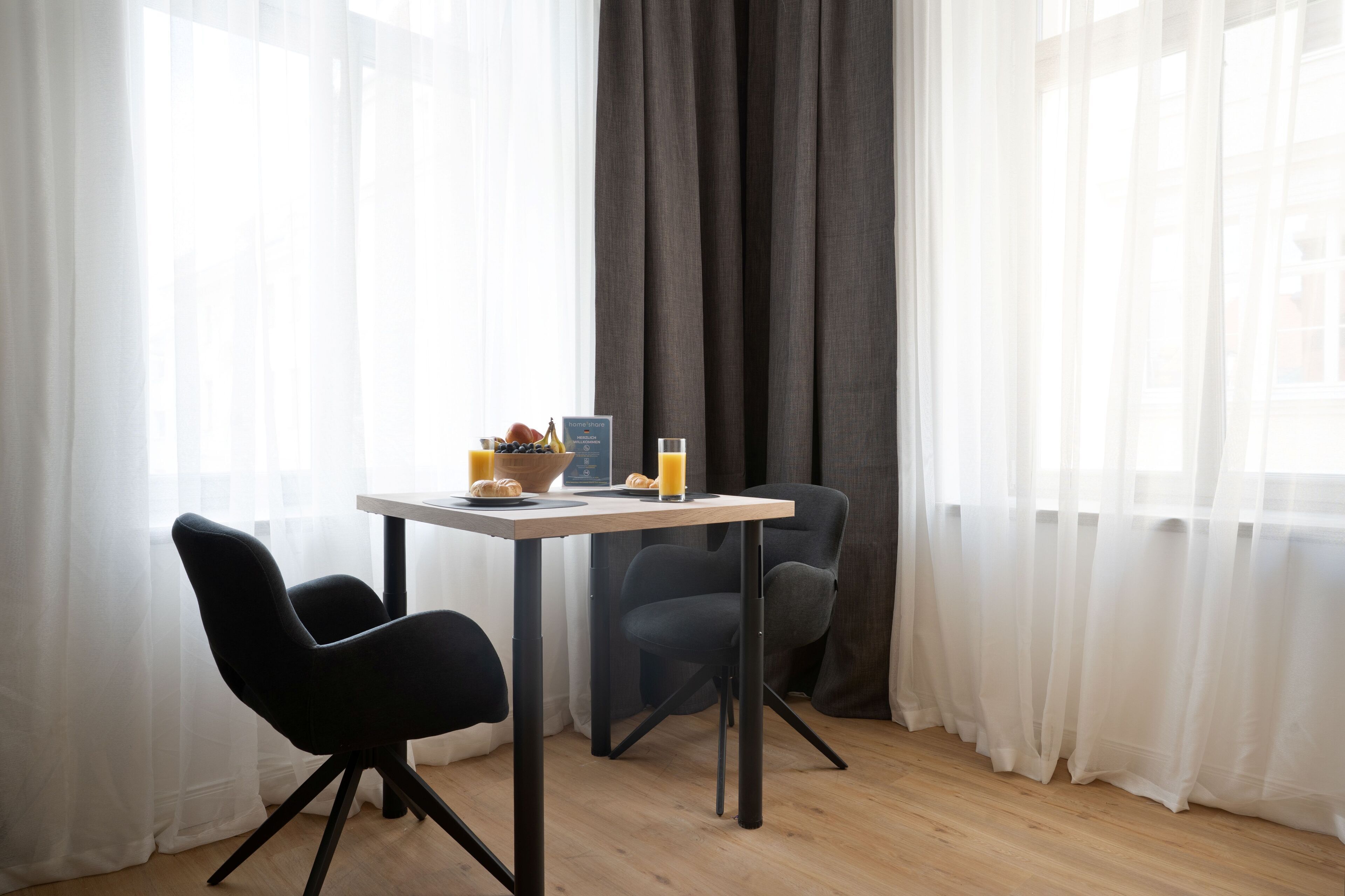 A modern room with a wooden floor, a table with breakfast items, two black chairs, and sheer curtains.