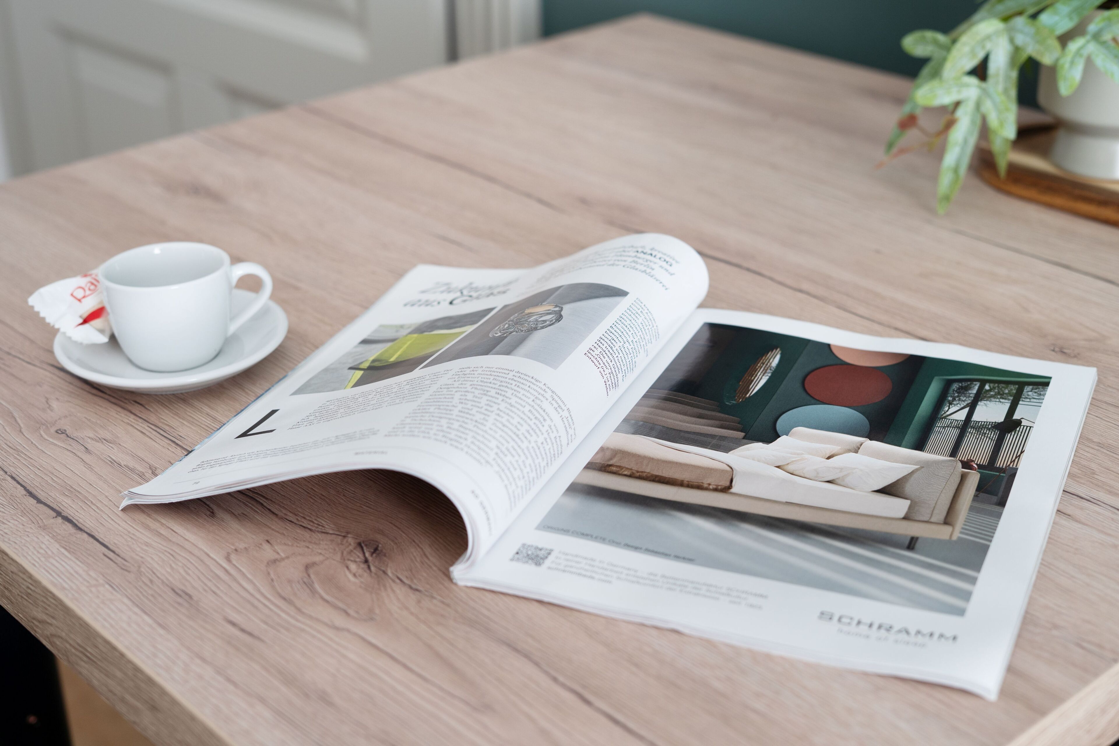 A magazine open on a wooden table, featuring a sofa and a cup with a saucer.